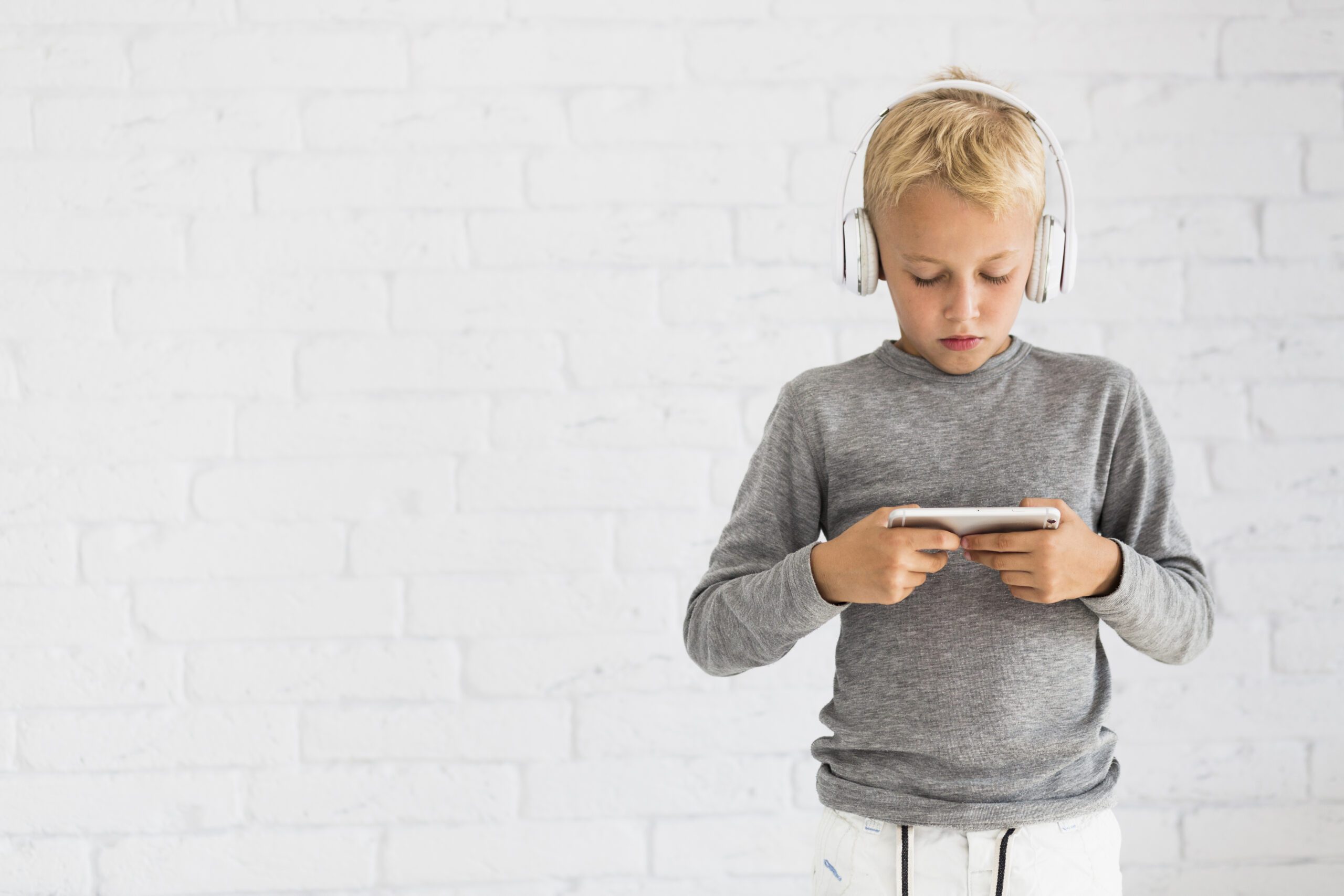 Young boy wearing headphones and focused on his smartphone, standing against a white brick wall.