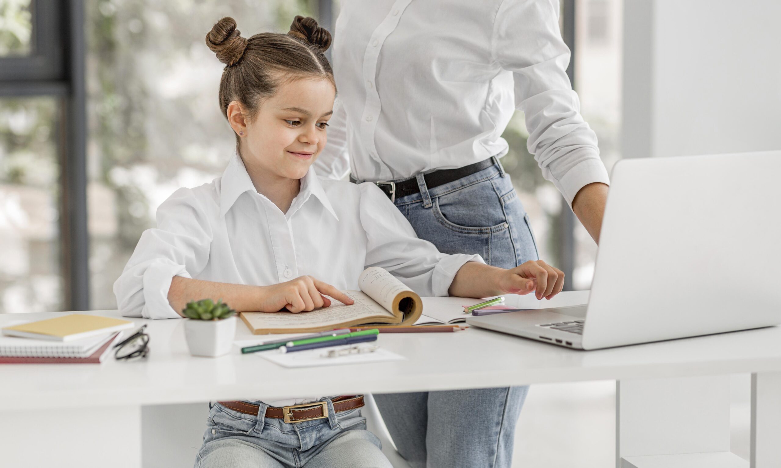 Young girl sitting at a desk with books and a laptop, guided by a parent.