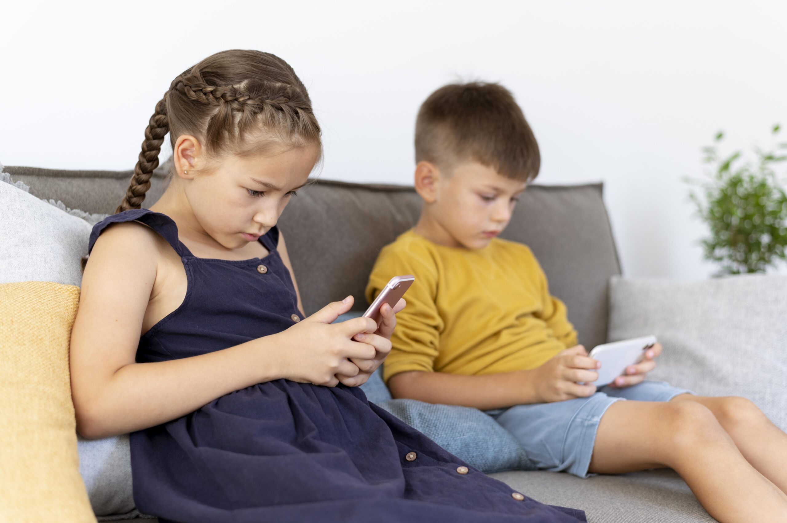 Two children sitting on a couch indoors, both focused on using their smartphones. Two children sitting on a couch indoors, both focused on using their smartphones.